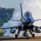 Blue fighter jet on runway with smoke behind it and a ground crew member in blue standing beside the aircraft cockpit canopy visible and orange chocks at the nose gear