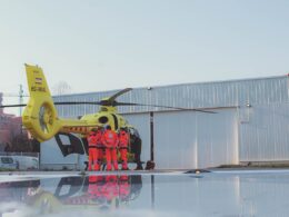 Yellow air ambulance helicopter on the tarmac with paramedics in red uniforms gathered by the open door of the cabin and a hangar in the background