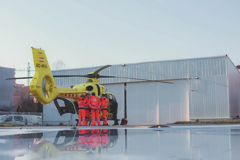 Yellow air ambulance helicopter on the tarmac with paramedics in red uniforms gathered by the open door of the cabin and a hangar in the background