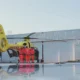 Yellow air ambulance helicopter on the tarmac with paramedics in red uniforms gathered by the open door of the cabin and a hangar in the background