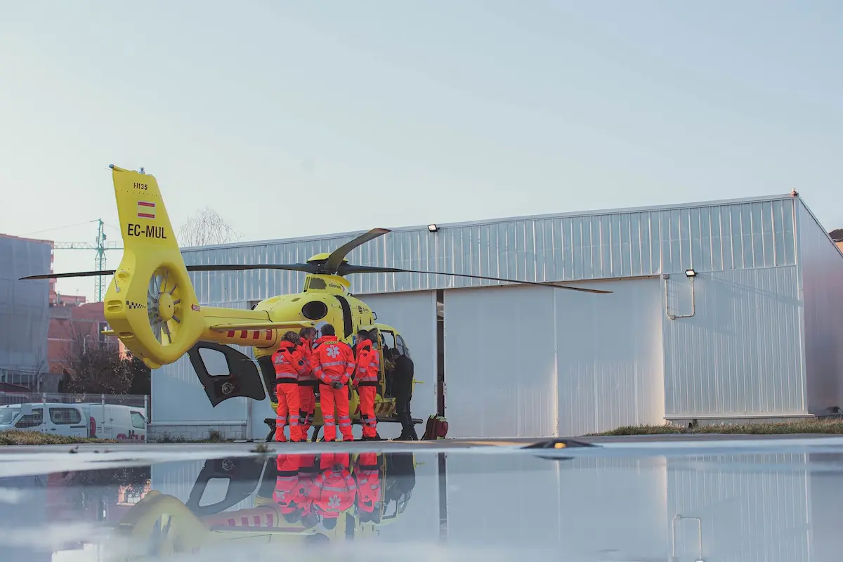 copilot pilot metge infermera hems sabadell metrosud xaviblanco img 8838 Yellow air ambulance helicopter on the tarmac with paramedics in red uniforms gathered by the open door of the cabin and a hangar in the background
