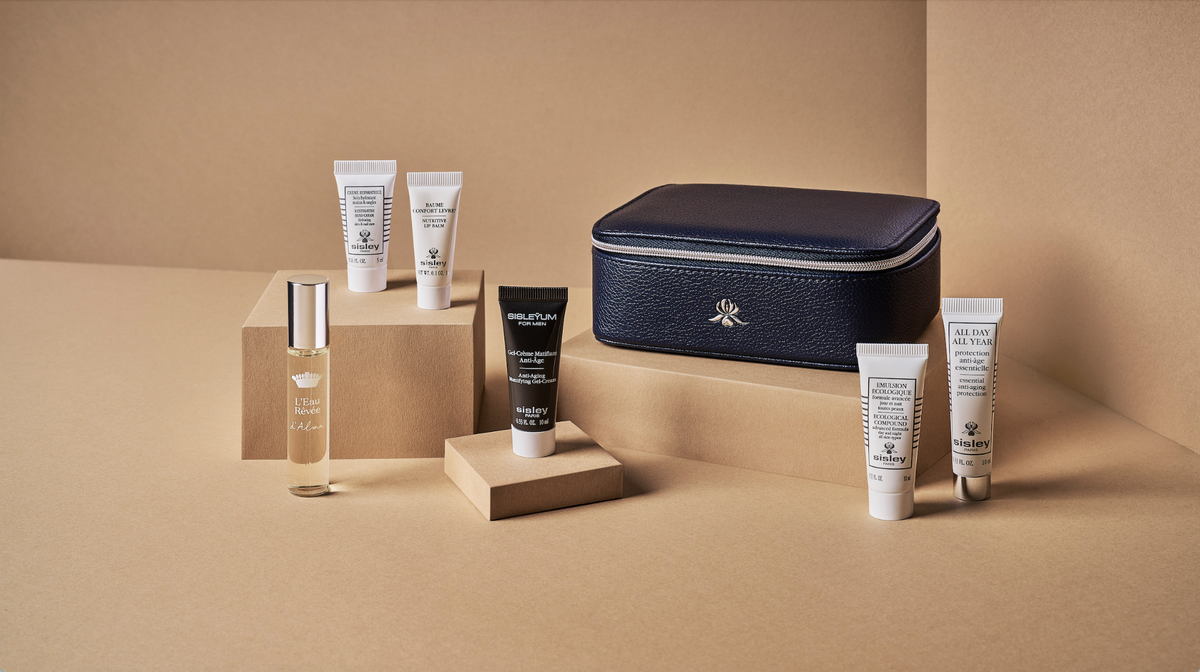 image Assorted skincare tubes a glass perfume bottle and a black sample tube arranged on beige display blocks beside a navy leather zippered case against a tan background