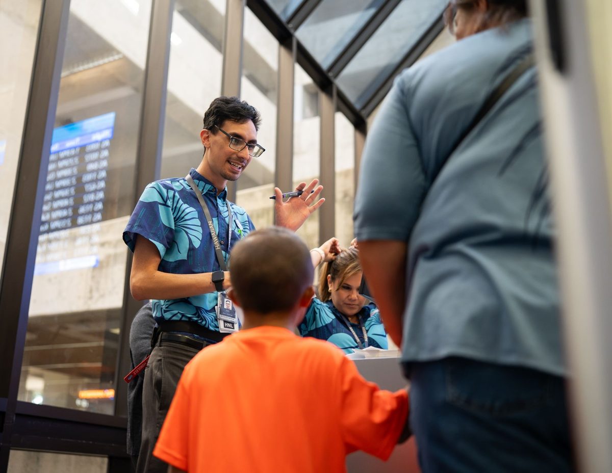 image Man with glasses in a blue patterned shirt hands a pen to children in orange shirts inside a glass walled lobby or atrium