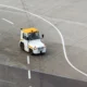 White service vehicle with a yellow roof driving on an empty concrete tarmac with curved white markings beside it