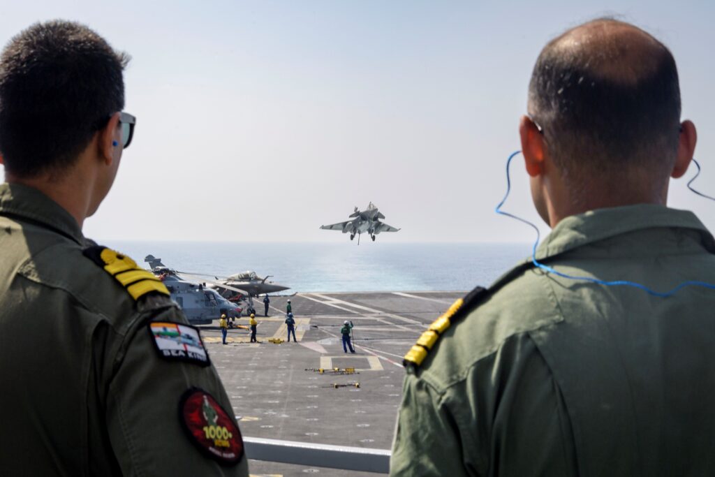 Two service members of the Indian Navy watch a Rafale fighter arrested landing on the Charles de Gaulle aircraft carrier