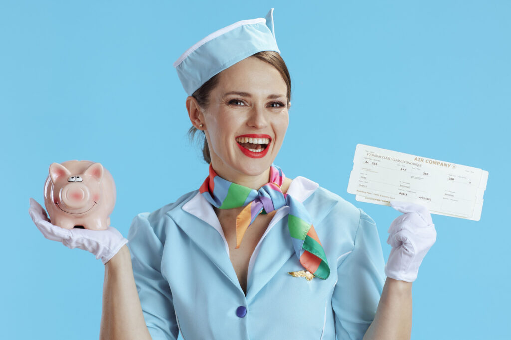 happy elegant female stewardess against blue background in blue uniform with flight tickets and piggy bank.